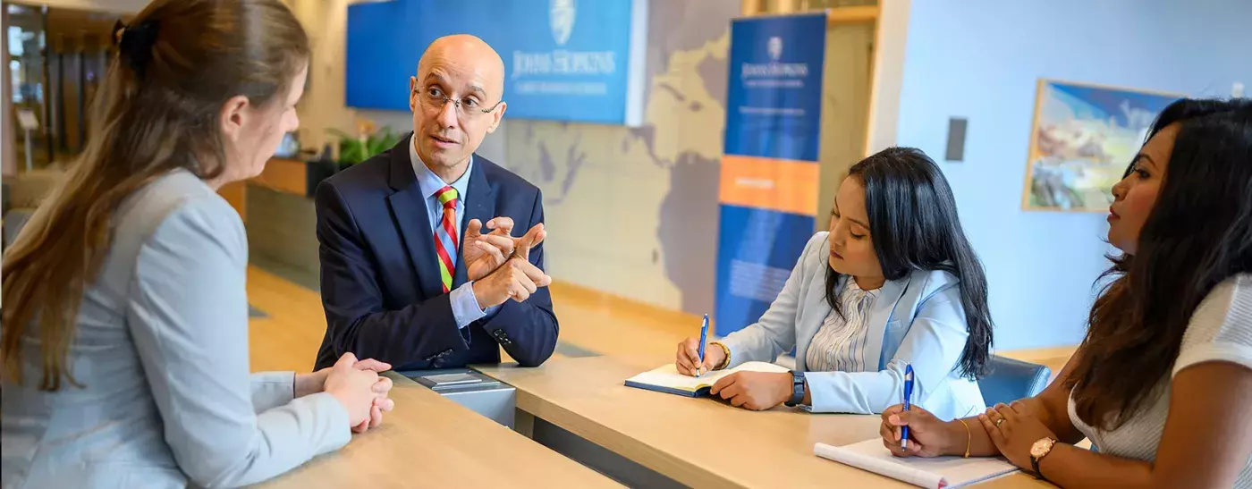 three students listening to a professor and taking notes sitting at a table