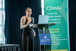 A woman stands behind a podium, with a green banner next to her, on the right side of the photo.