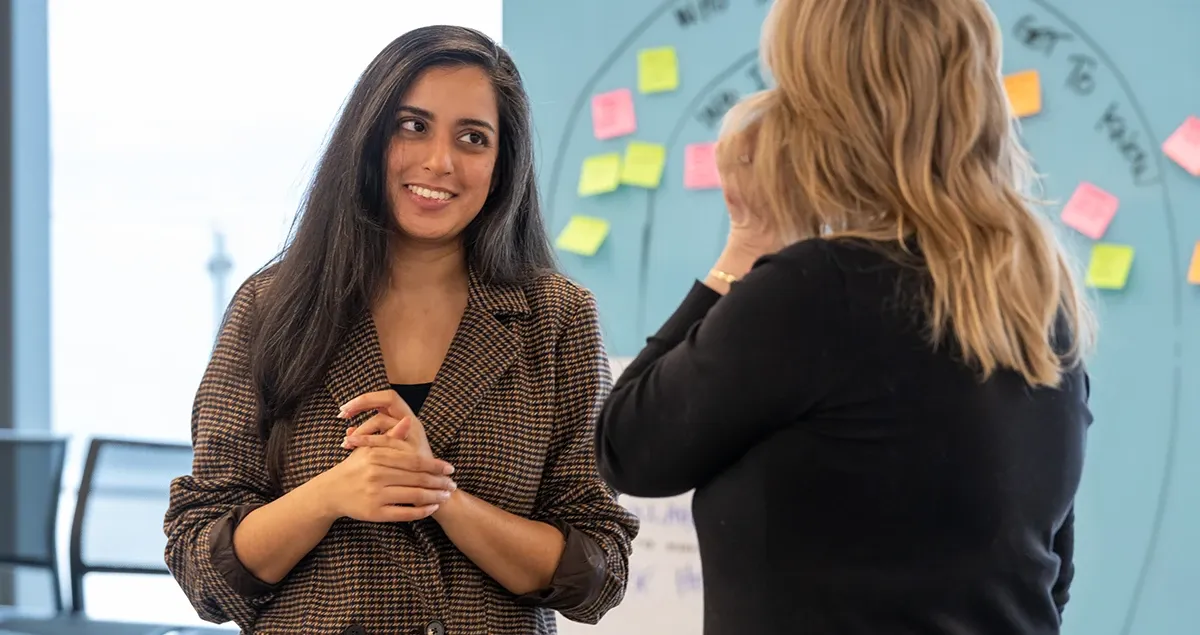 Two women speak in a professional setting during a leadership coaching or workshop session, with sticky notes on a glass board in the background.