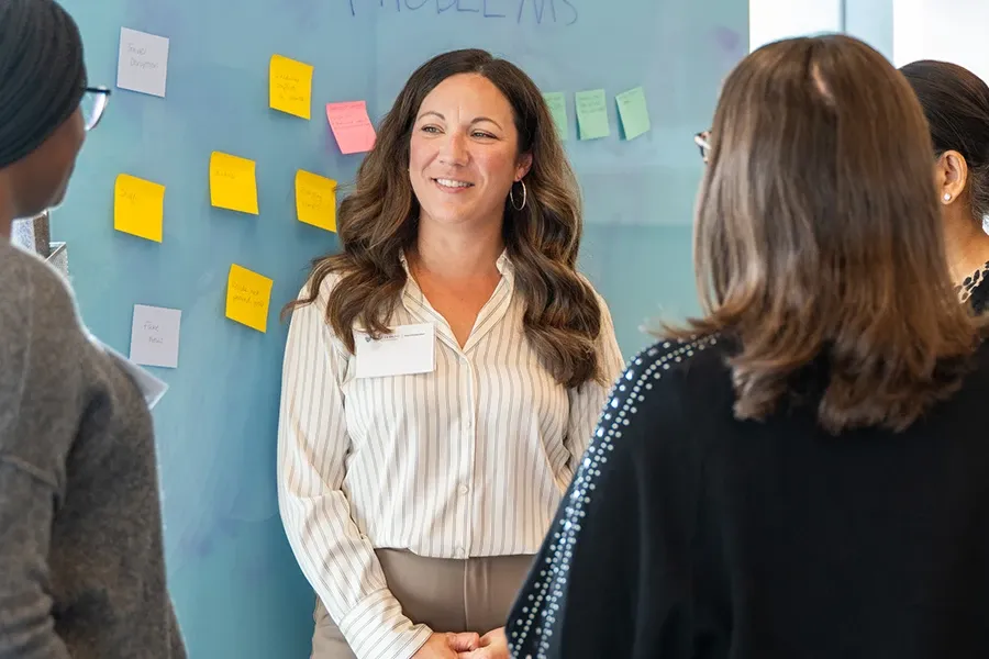 Participants gather around a glass wall covered with colorful sticky notes during a design thinking workshop, as a facilitator leads a collaborative discussion