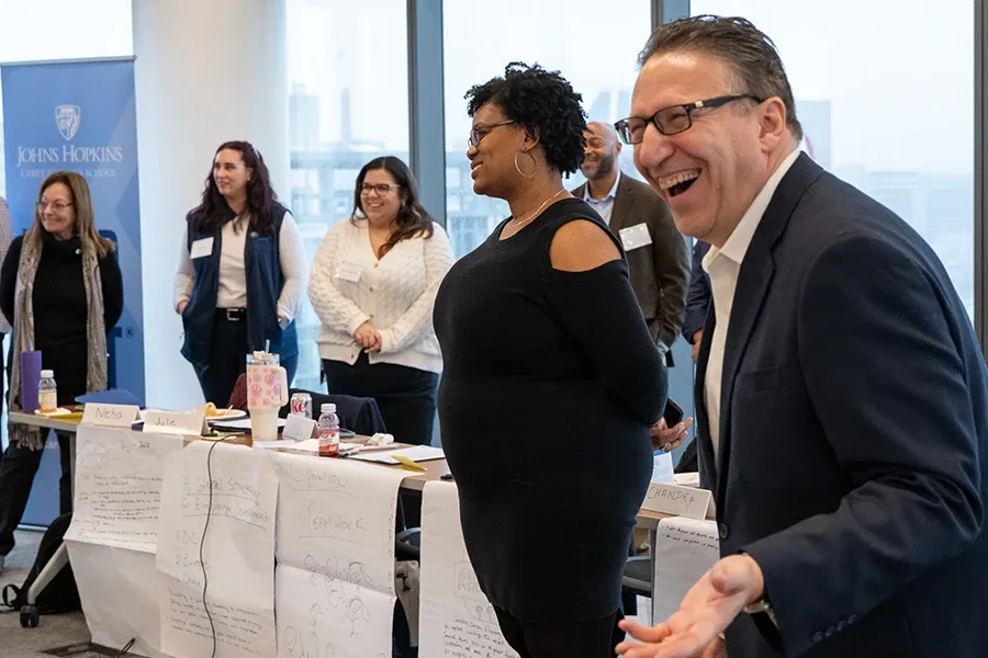 Workshop participants stand and engage in a group facilitation exercise in a classroom setting, with brainstorming materials displayed around the room and a participant laughing during the activity