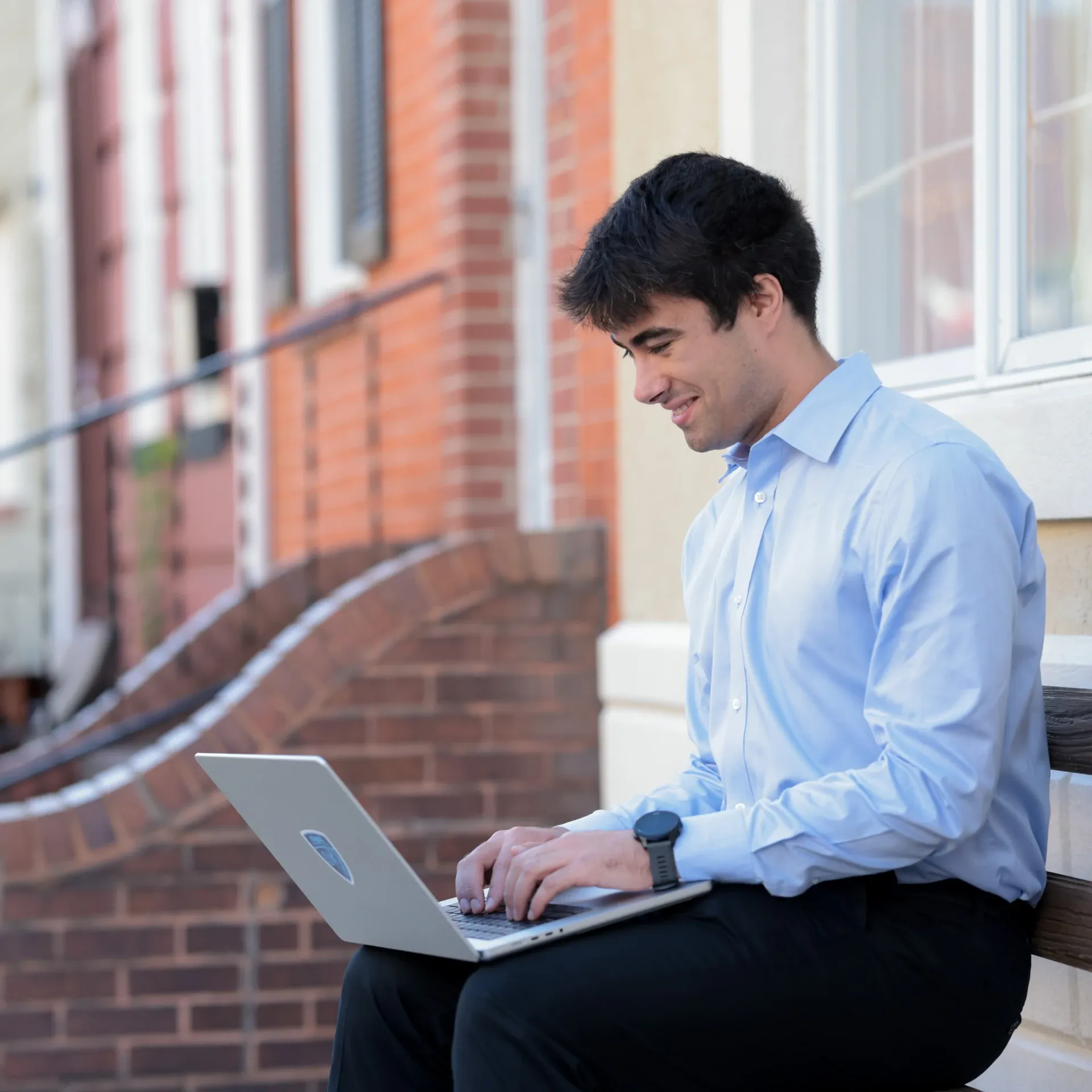 Man in blue shirt sitting on steps, typing on laptop outside a brick building
