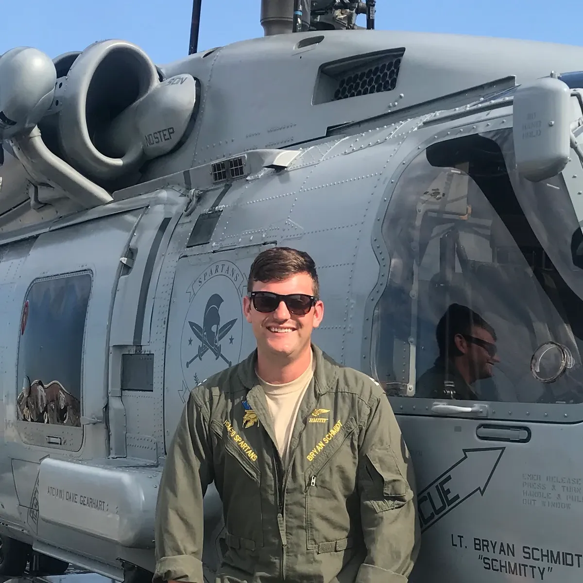 Bryan Schmidt in flight suit and sunglasses in front of a gray military helicopter.