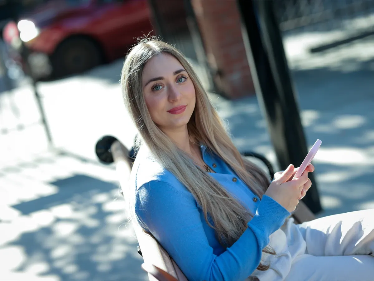 Smiling blonde woman in blue blazer holding a smartphone outdoors on a sunny day