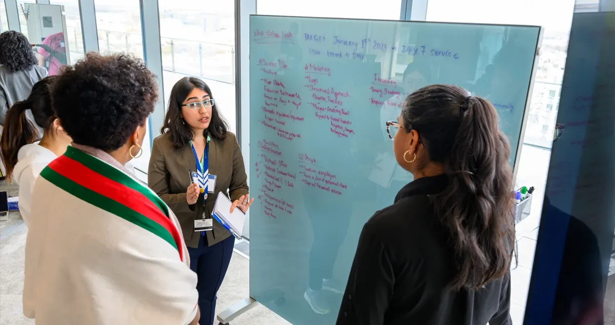 Attendees discuss notes on a large glass whiteboard in a bright room.