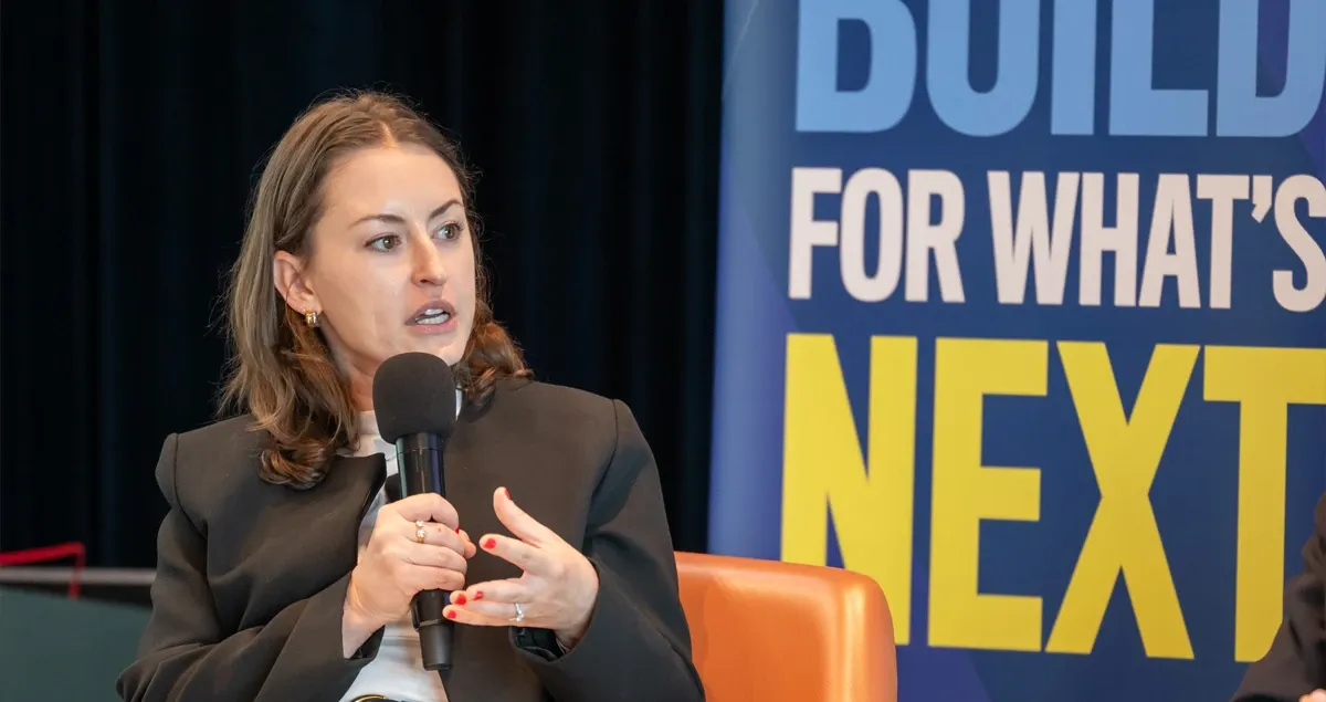 Woman speaking into a microphone onstage beside “BUILD FOR WHAT’S NEXT” banner
