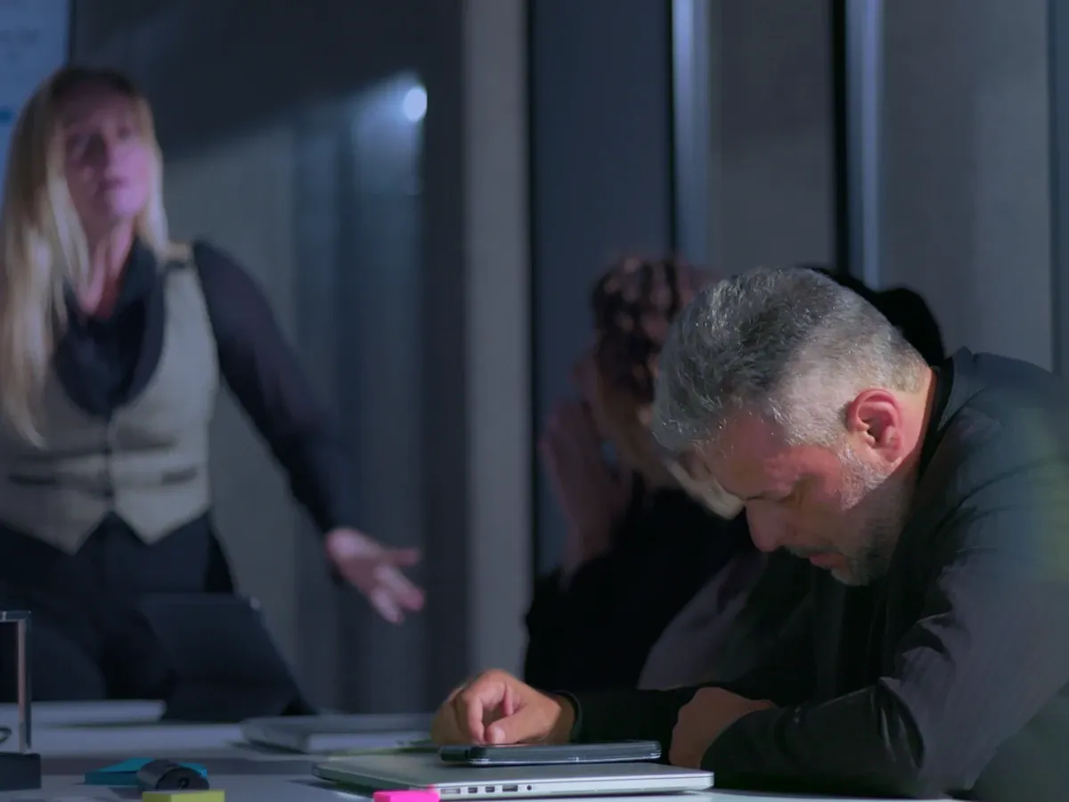 Man slumped at desk, head down, as blurred woman gestures in background.