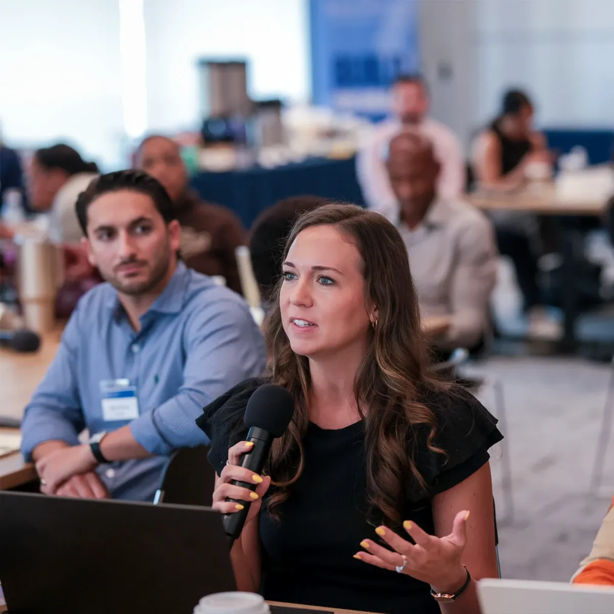 Woman speaking into a microphone during a meeting, with attendees blurred in the background