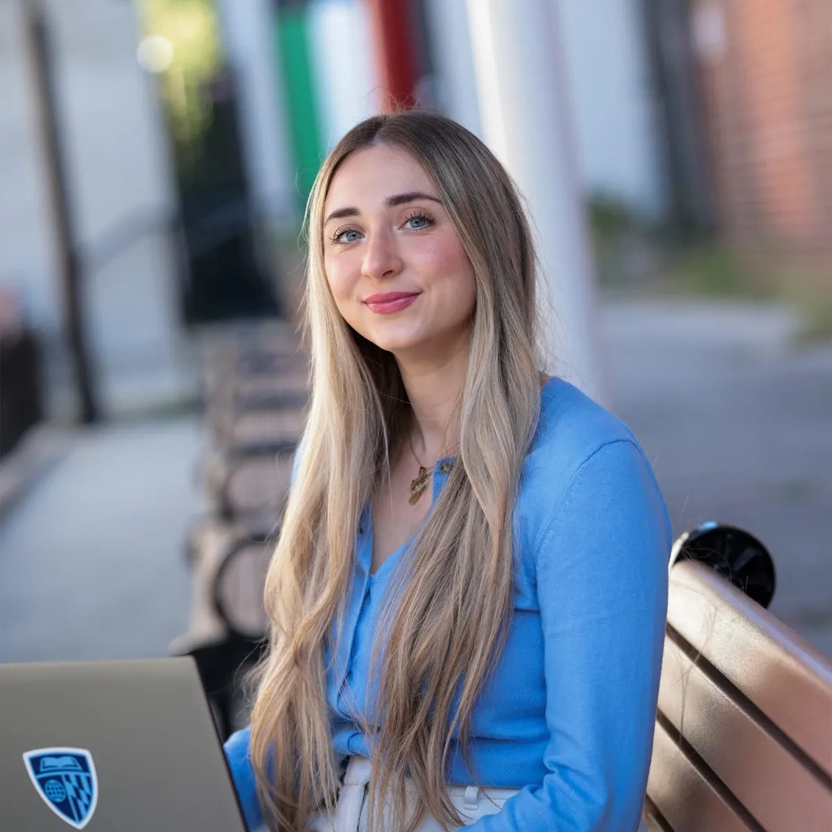 Smiling woman in a blue top sitting on a bench with an open laptop outdoors