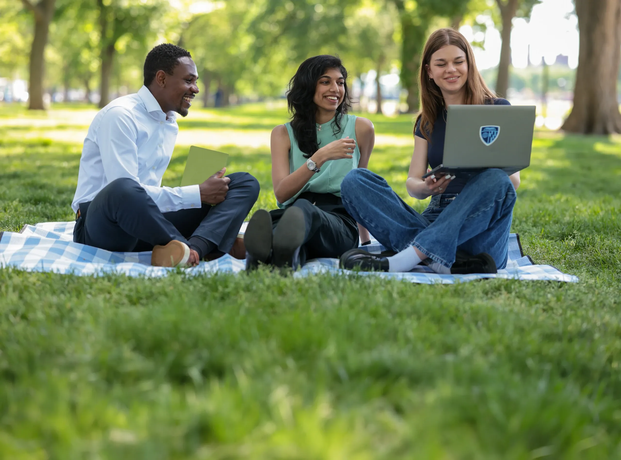 Three students on a picnic blanket in a park, smiling with laptops