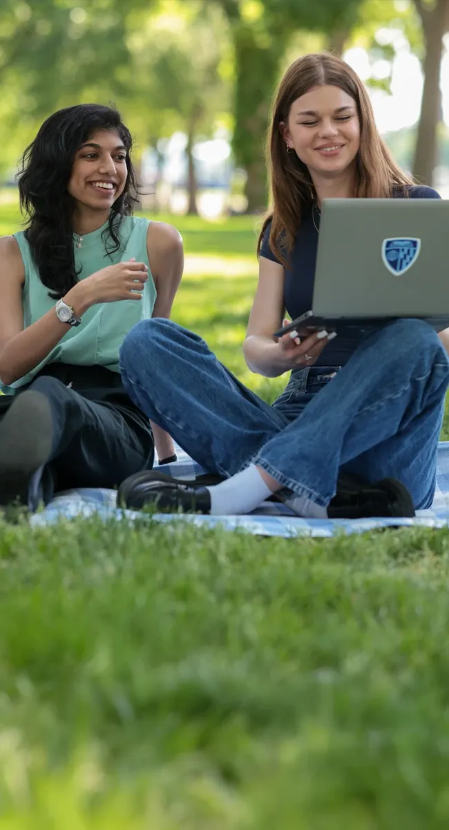Two students sitting on grass in a park, smiling beside an open laptop