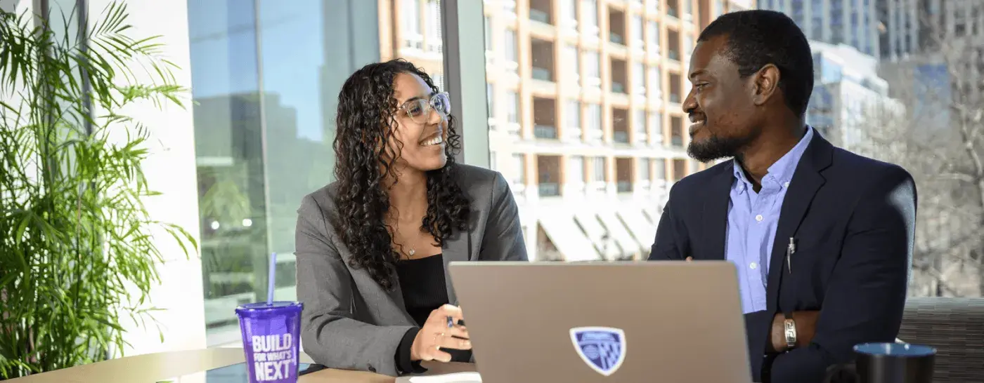 Two professionals in a modern office setting having a conversation at a table, smiling and seated with a laptop and Carey-branded tumbler.