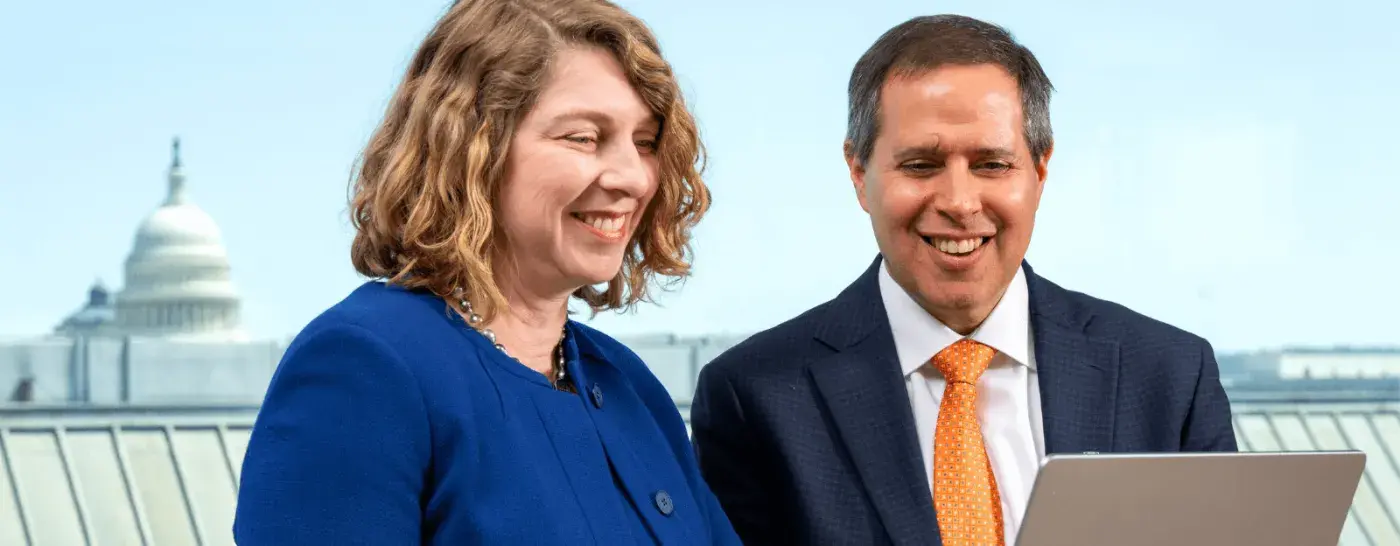 Two business professionals smiling and looking at a laptop together, with the U.S. Capitol building visible in the background through a window.