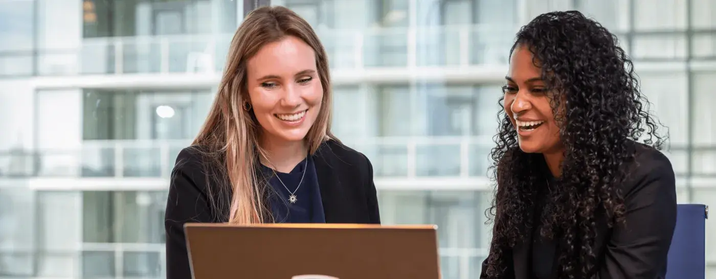Two women in professional attire smiling and collaborating over a laptop, with tall glass windows in the background.