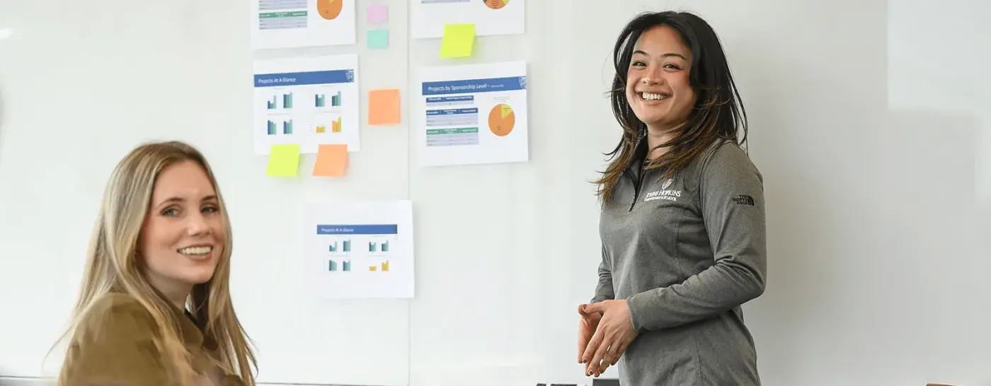 Two women engaged in a discussion in front of a whiteboard covered with charts and sticky notes, one standing and smiling while the other is seated and turning toward the camera.