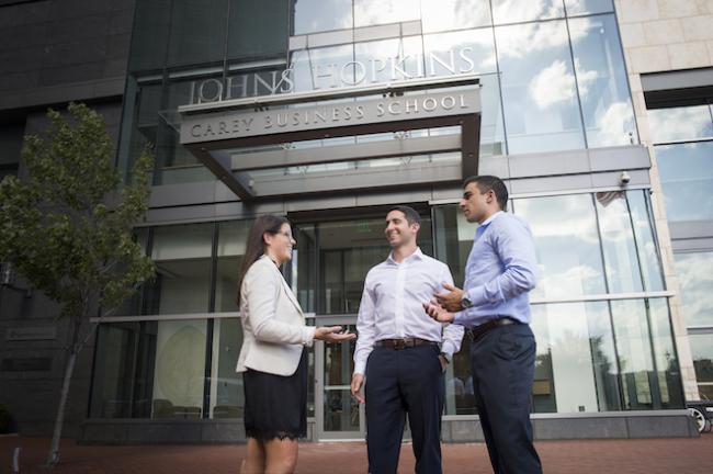 Three students stand in front of the entrance to the Johns Hopkins Carey Business School