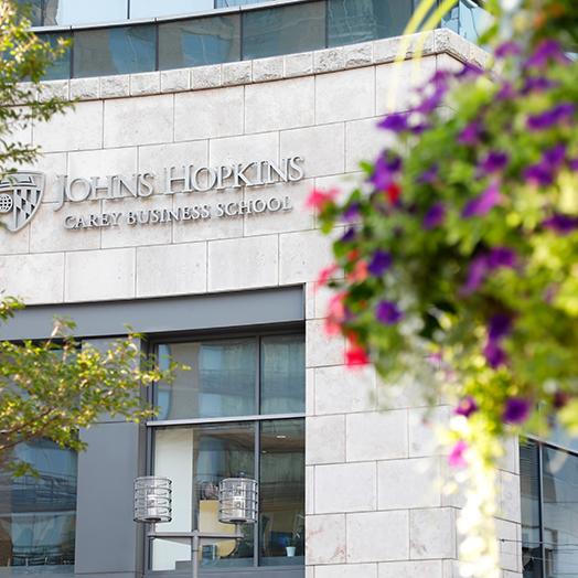 Entrance Johns Hopkins Carey Business School during the day with logo facade and tree limbs and hanging flowers