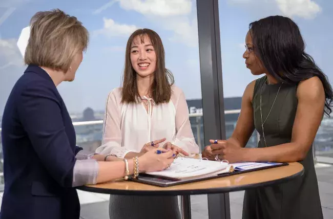 3 students standing around a circular table discussing an open notebook
