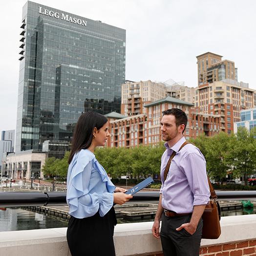 a couple of students outside on a foot bridge over looking the Baltimore buildings and holding a Carey Business school folde