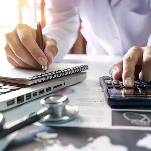 stock image of health professional with pen writing in a notebook with one hand and the other entering numbers into a calculator.