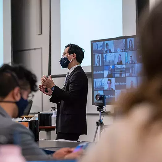 professor speaking with a mask to in person masks students with a zoom students screens in background