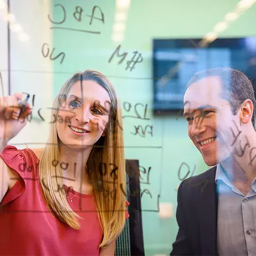 2 people as seen from behind a transparent white board with a dry erase calculating  ABC table