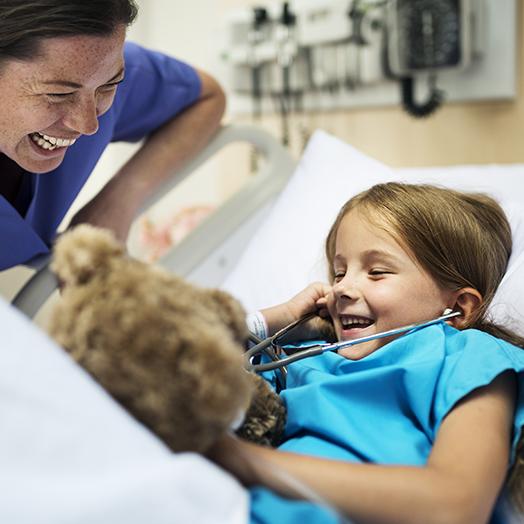 small child in a hospital bed with a teddy bear and a medical staff leaning over and smiling