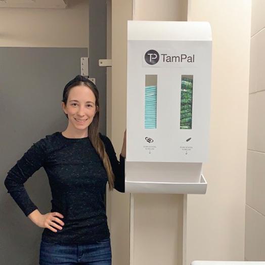 Erica Duffy standing beside her product a TamPal dispenser in a public restroom