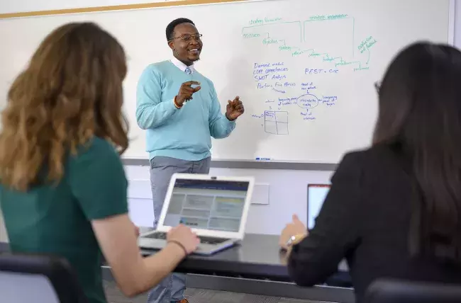 instructor in front of a white board with 2 students seated taking notes on laptops 