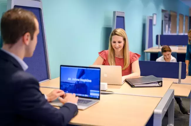 two people sitting at ajoining tables with laptops and people to the rear in a study area