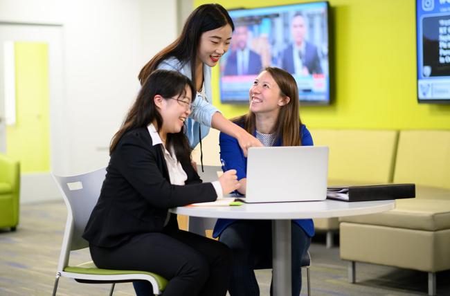 three students meeting over a laptop at a small round table. two are seated while the third is standing and pointing to something displayed not shown. tags: MBA Healthcare management, Online Healthcare MBA Program