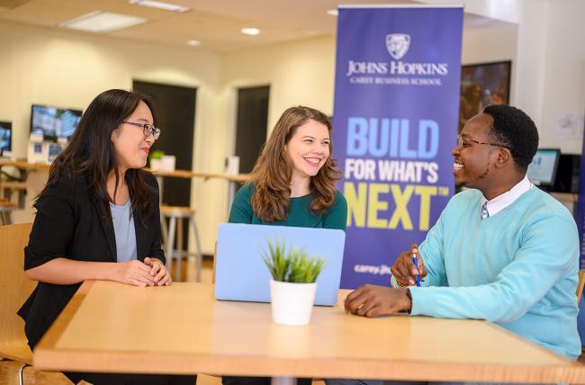three students sitting at a small square table with a laptop in a JHU Carey room that have a banner that reads Johns Hopkins Carey School of business logo with the Build for What's Next slogan
