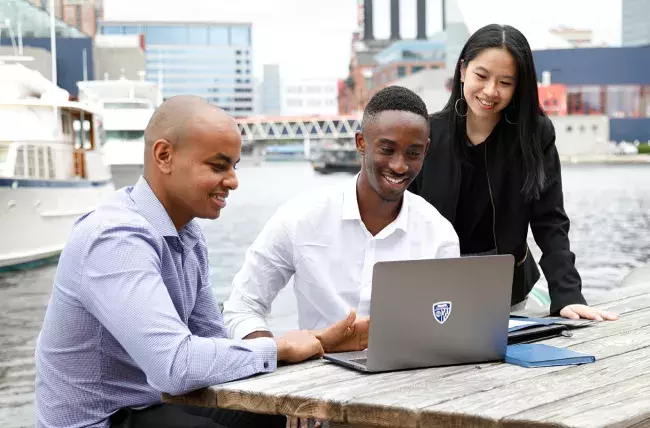 three students sharing a laptop on a picnic table by the harbor