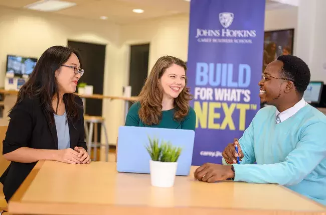 three students sitting at a table with build for what's next banner in background