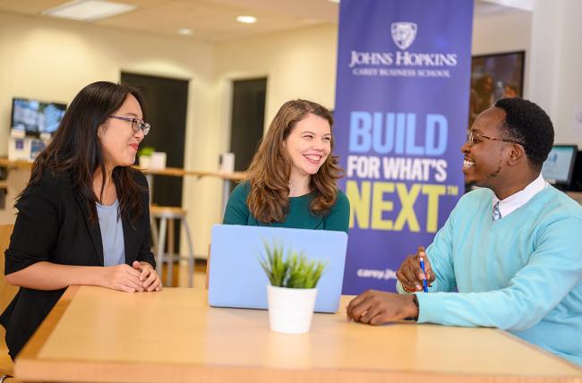 three students sitting at a table with build for what's next banner in background