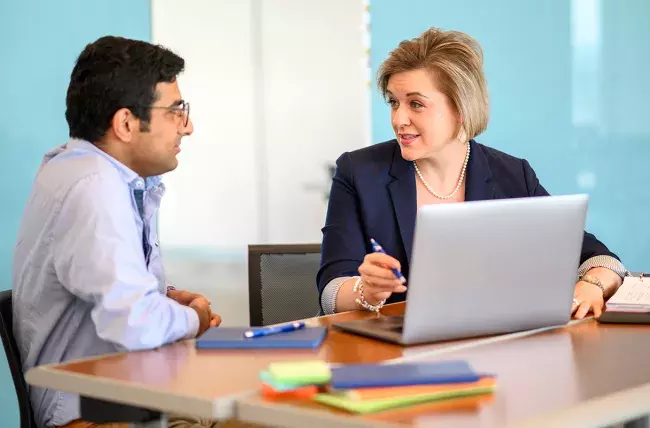 two people sitting at a table with one pointing to a laptop with a pen explaining 