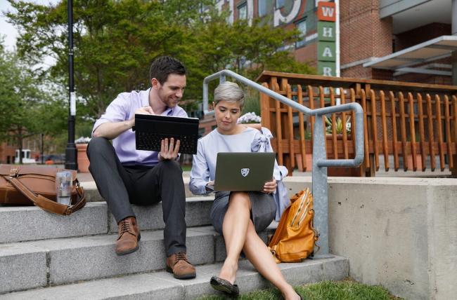 two students sitting casually on stone steps outside a building on campus comparing laptops displays