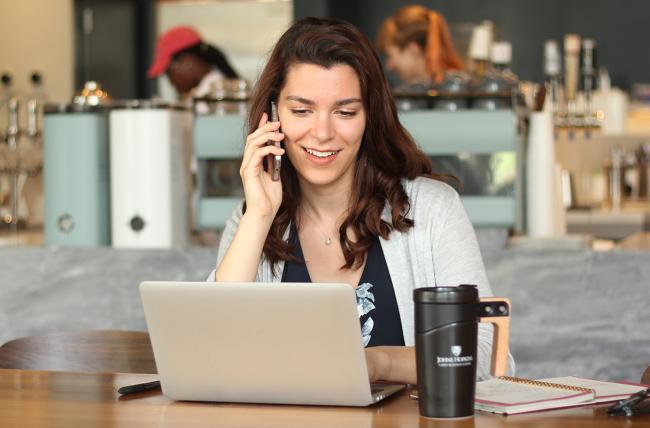 woman sitting at a table in a bistro talking on a mobile phone while working in a laptop with a thermos and notebook next to her 