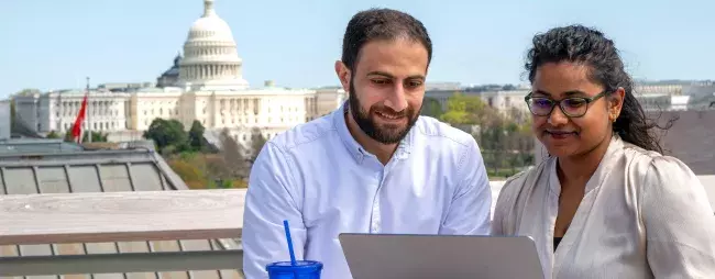 two students sharing a laptop no the top of the building with the capital in the background.