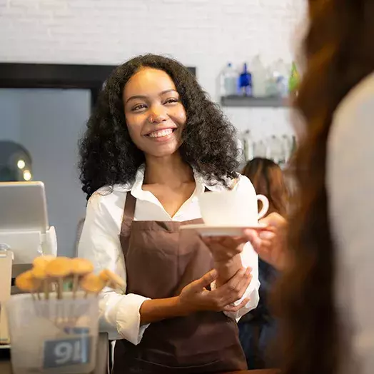 Female barista with black hair and a big smile offering a cup to a female customer