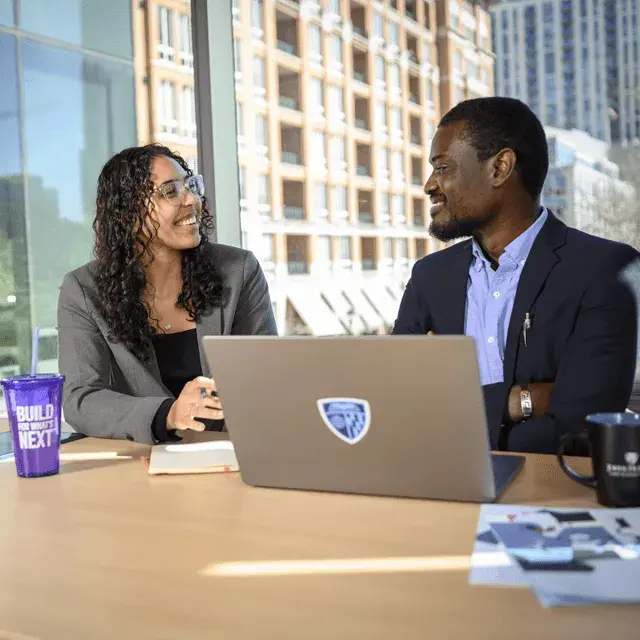 Two professionals smiling and talking at a table with a laptop and notebooks in a bright office setting