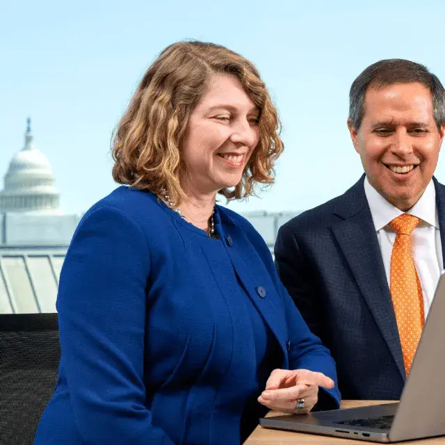 Two professionals smiling at a laptop with the U.S. Capitol in the background