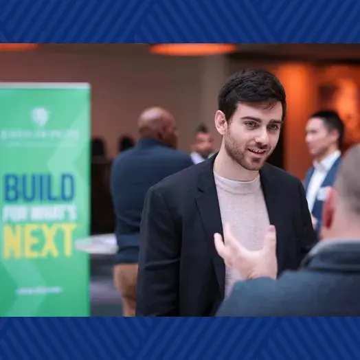 graduate student standing and listening to the a person seated who is the authority with others in the background and a Build for whats next sign