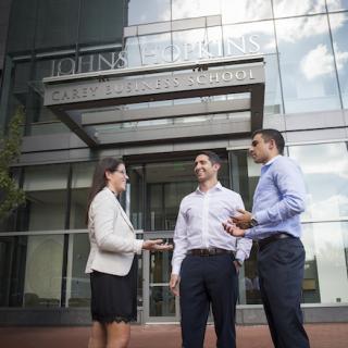 Three students stand in front of the entrance to the Johns Hopkins Carey Business School