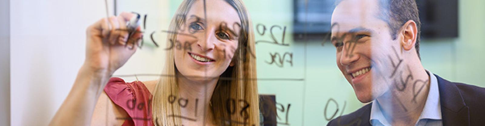 2 business professionals writing formulas on the otherside of a glass board