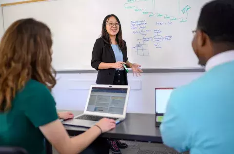 two students sitting at a table with another student demonstrating commercialization on a dry erase board