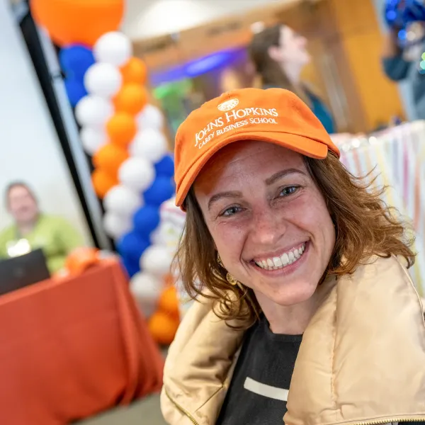 Smiling woman in orange cap and beige jacket at an indoor event with balloons