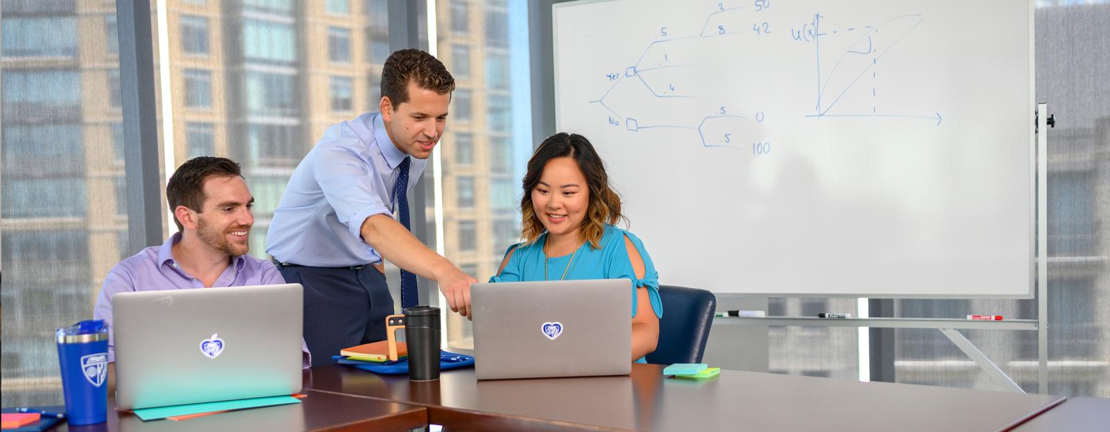 2 students at a table in a window filled meeting room with an instructor pointing to one of the laptops with a whiteboard in the back that has a decision tree and a x/y curve