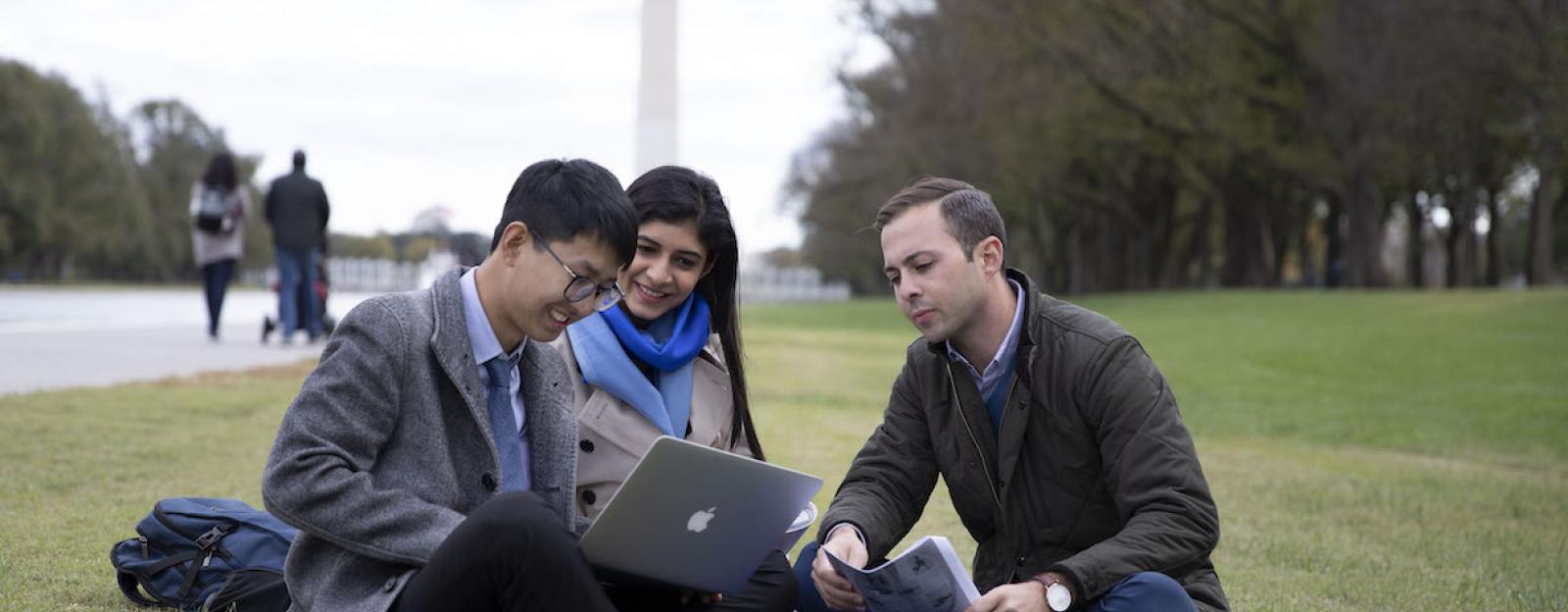 3 students siting on lawn in front of the Washington monument.