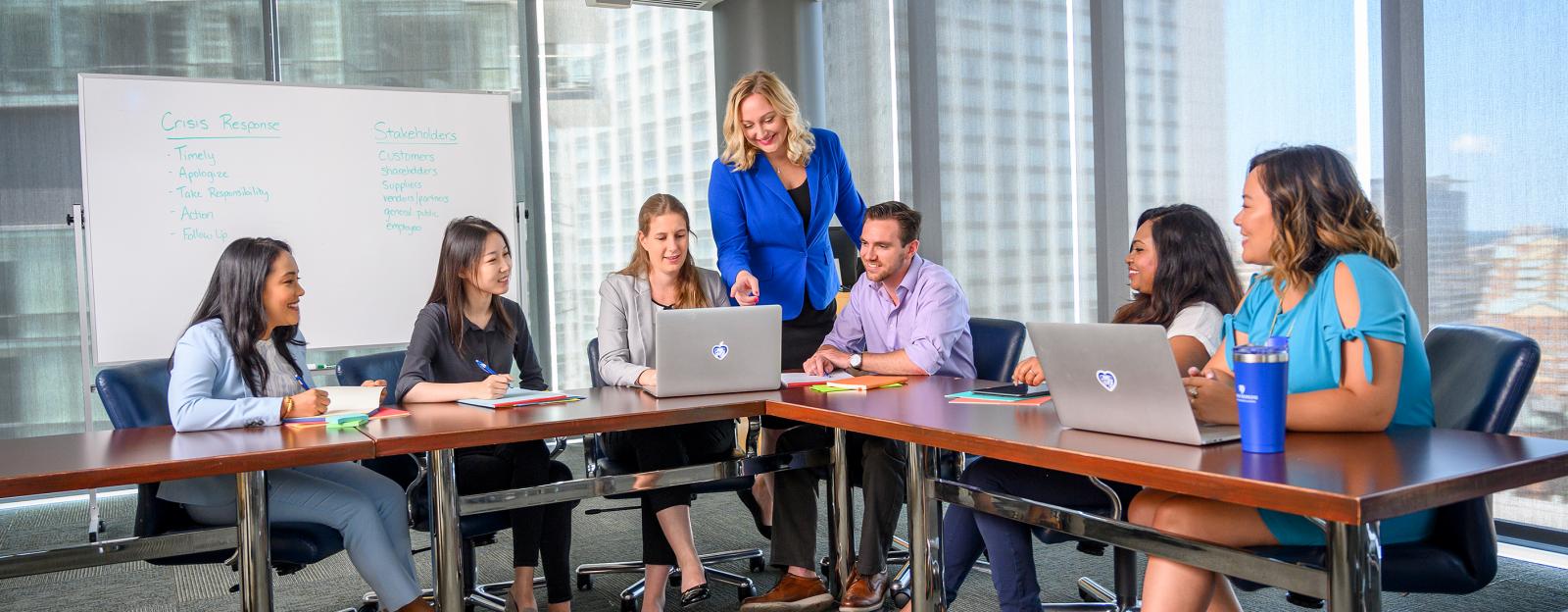 6 students taking notes seated at a table taking notes while an instructor points to a laptop